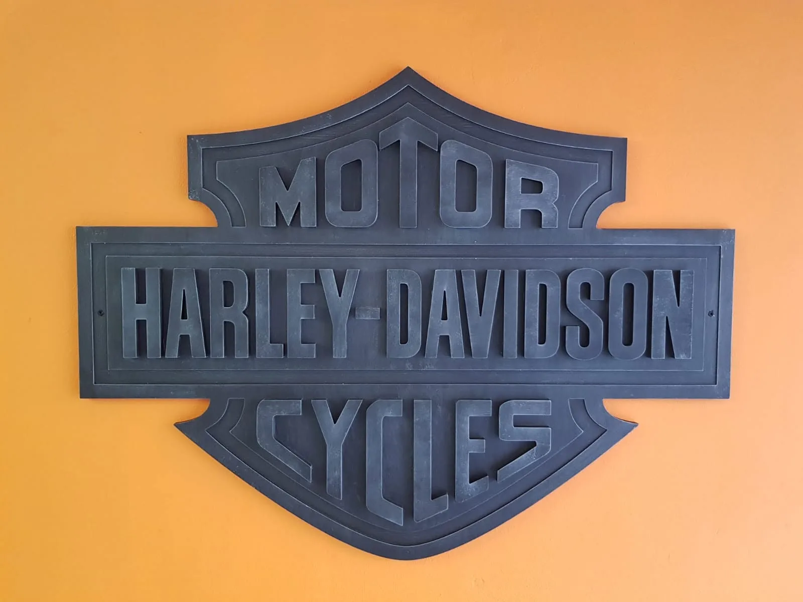 Čierne vrstvené priestorové logo Harley-Davidson vyfrézované na CNC a osadené na oranžovej stene