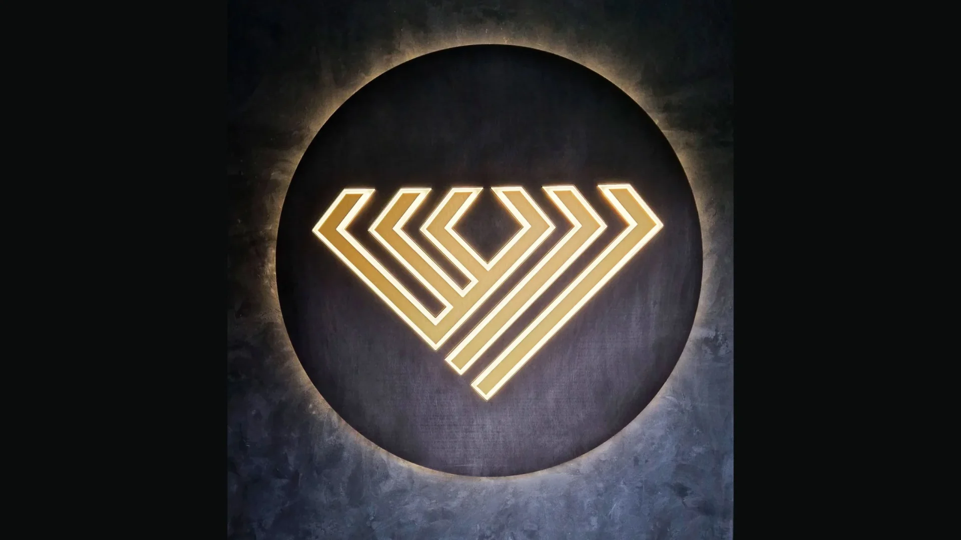 Zlaté geometrické logo na tmavom kruhovom podklade s halo podsvietením