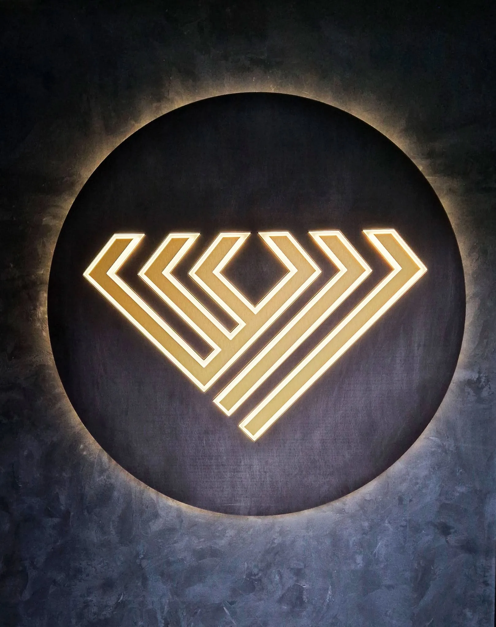 Zlaté geometrické logo na tmavom kruhovom podklade s teplým halo podsvietením zozadu