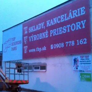 Reklamný banner s ponukou skladov a kancelárií na stene priemyselnej haly, montáž z vysokozdvižnej plošiny