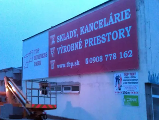 Reklamný banner s ponukou skladov a kancelárií na stene priemyselnej haly, montáž z vysokozdvižnej plošiny