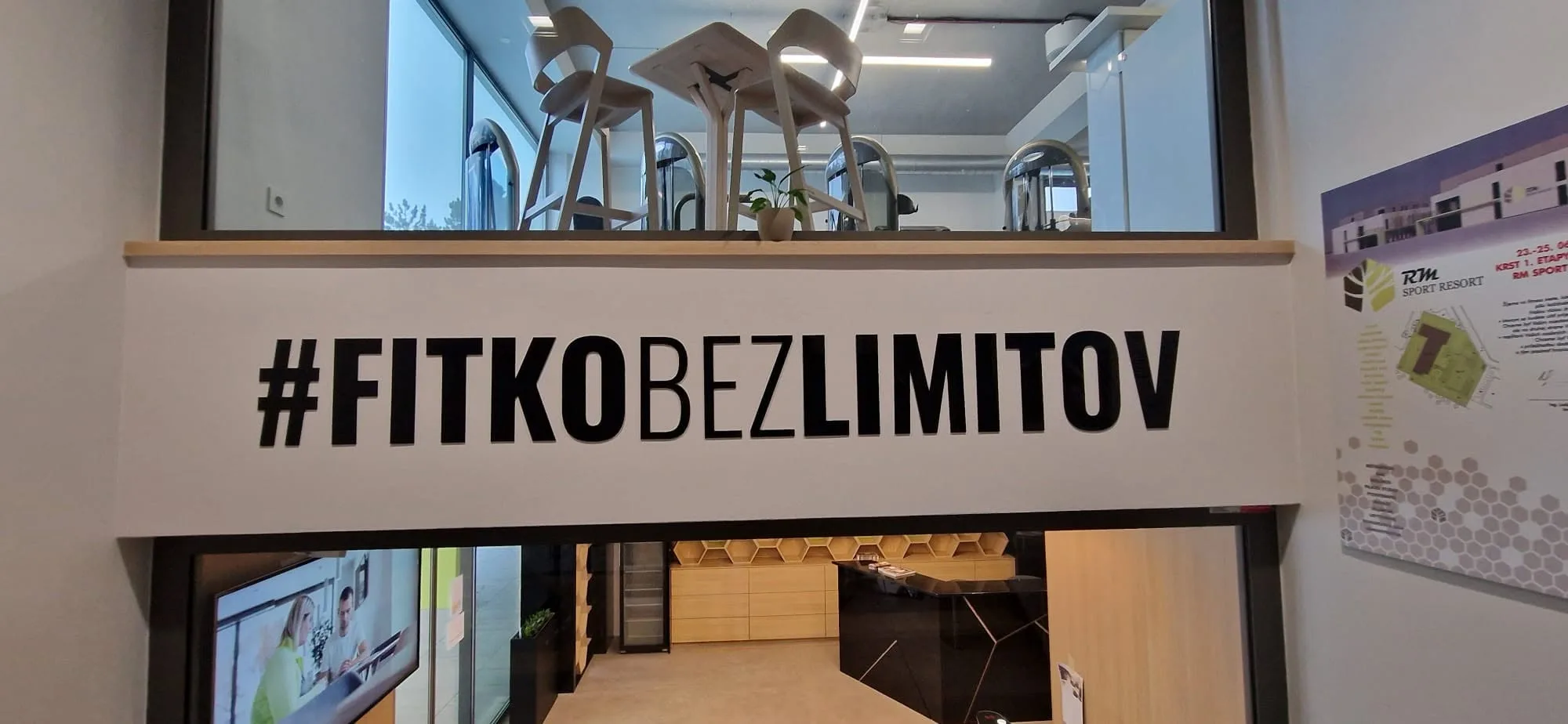 Čierny priestorový nápis #FITKOBEZLIMITOV nad presklenou stenou fitnescentra