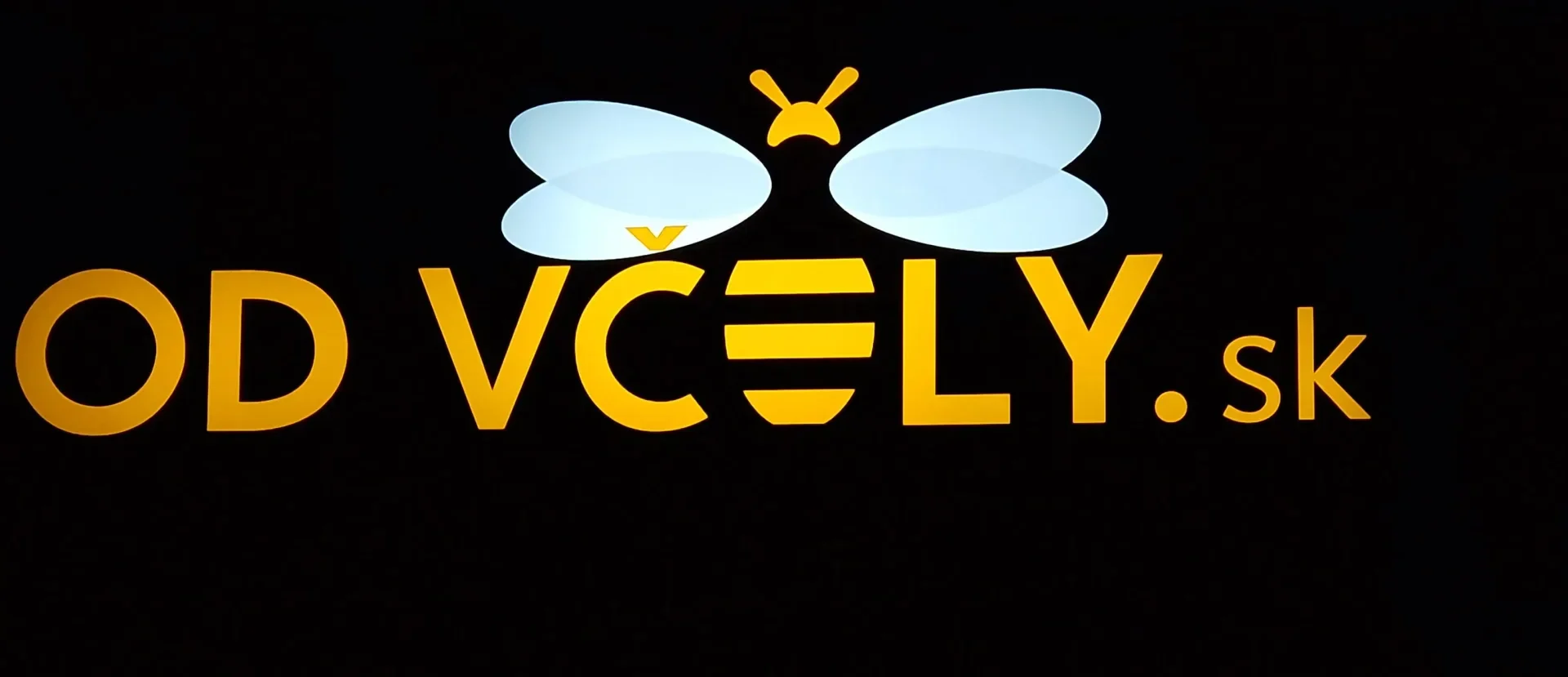 Rozsvietený nápis Od včely.sk s logom včely, žltý text a biele krídla žiaria v tme