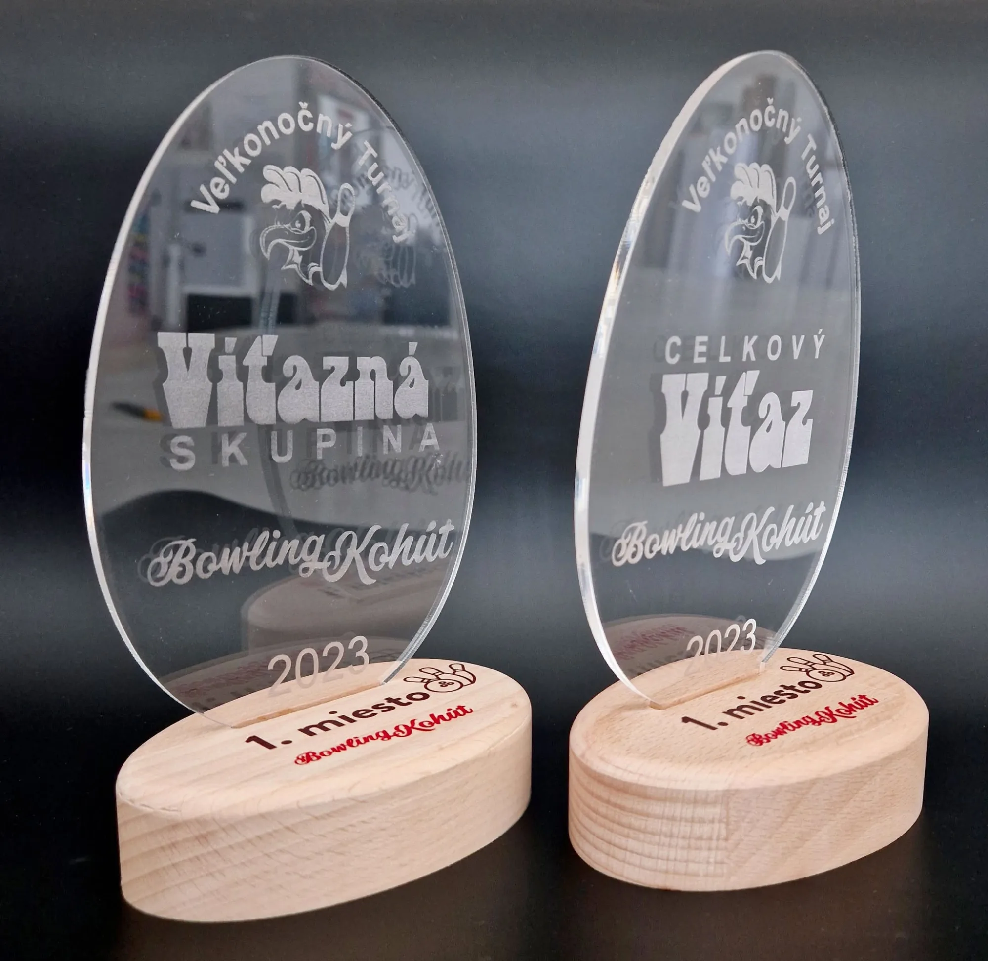 Dve trofeje v tvare vajca z číreho akrylátu s laserovým gravírom na drevených podstavcoch
